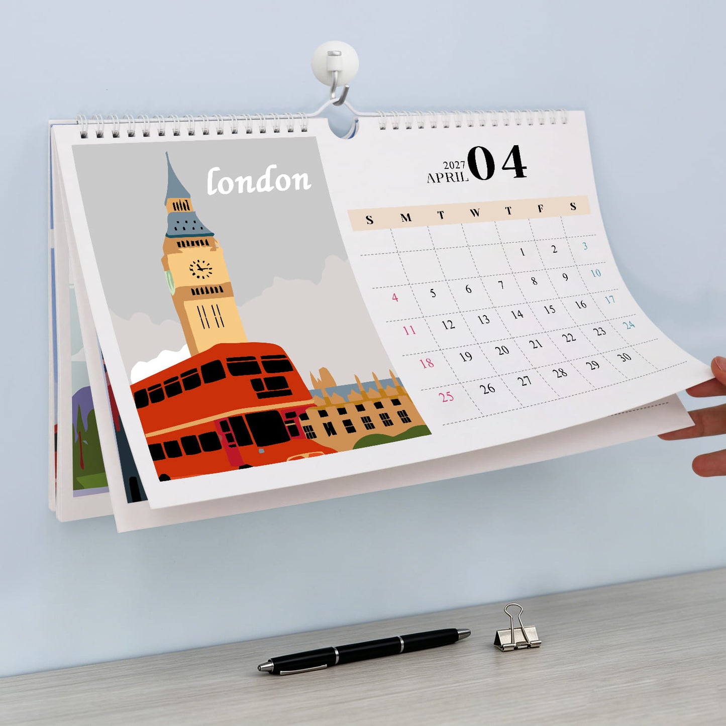 A spiral-bound desk calendar showing London, with a hand holding the April page open against a light blue wall above a desk with a pen and binder clip.