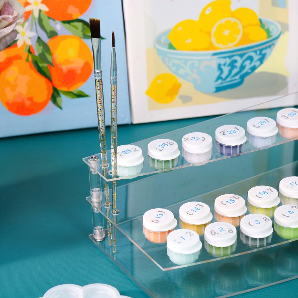 Set of paint pots with brushes on a teal surface, with a painting of lemons in the background.