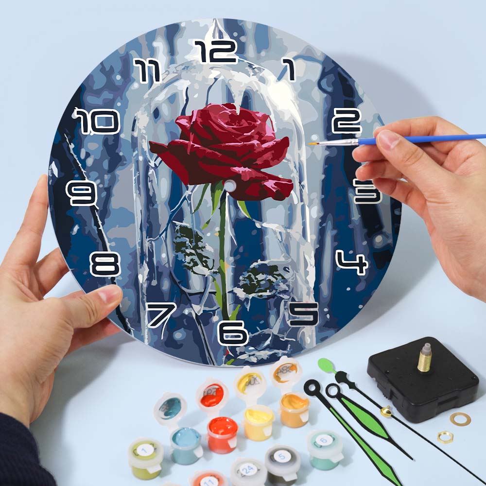 Person painting a rose on a round clock face with paint supplies on a table.