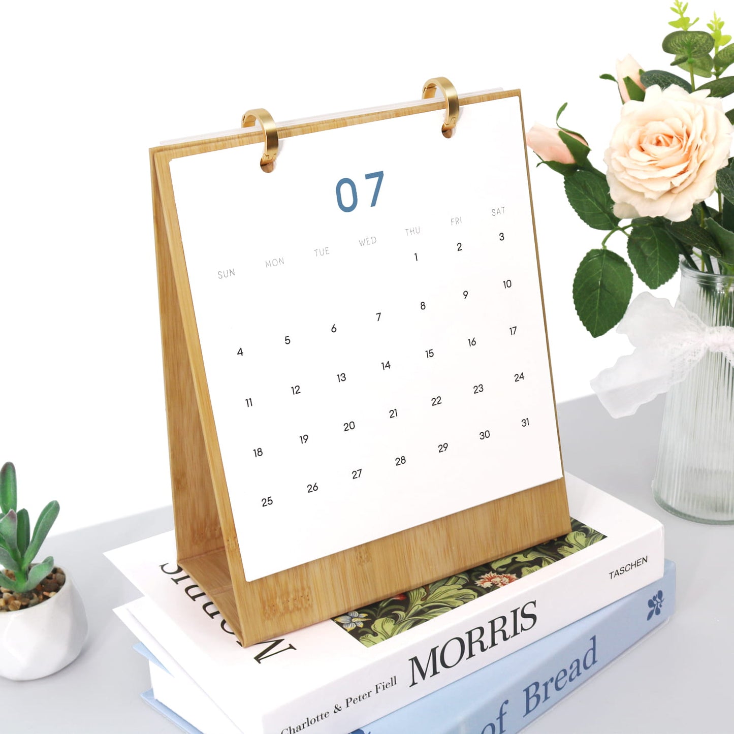 A bamboo desktop calendar stand holding a white July page, styled on top of books with a small plant and flowers in the background.