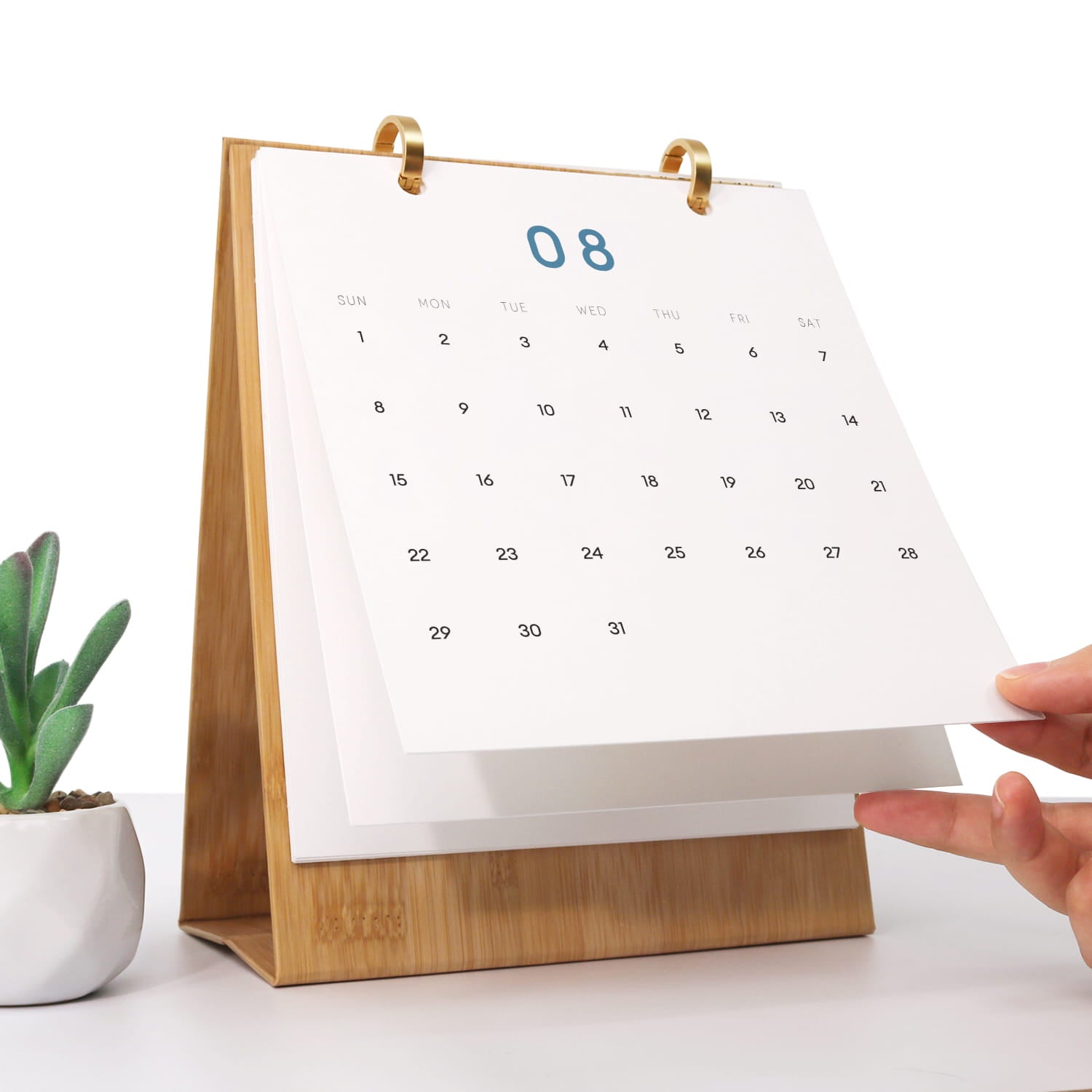 A bamboo desktop calendar with gold metal rings holding white pages, shown on a white surface with a small potted plant at left and a hand holding one page at right.