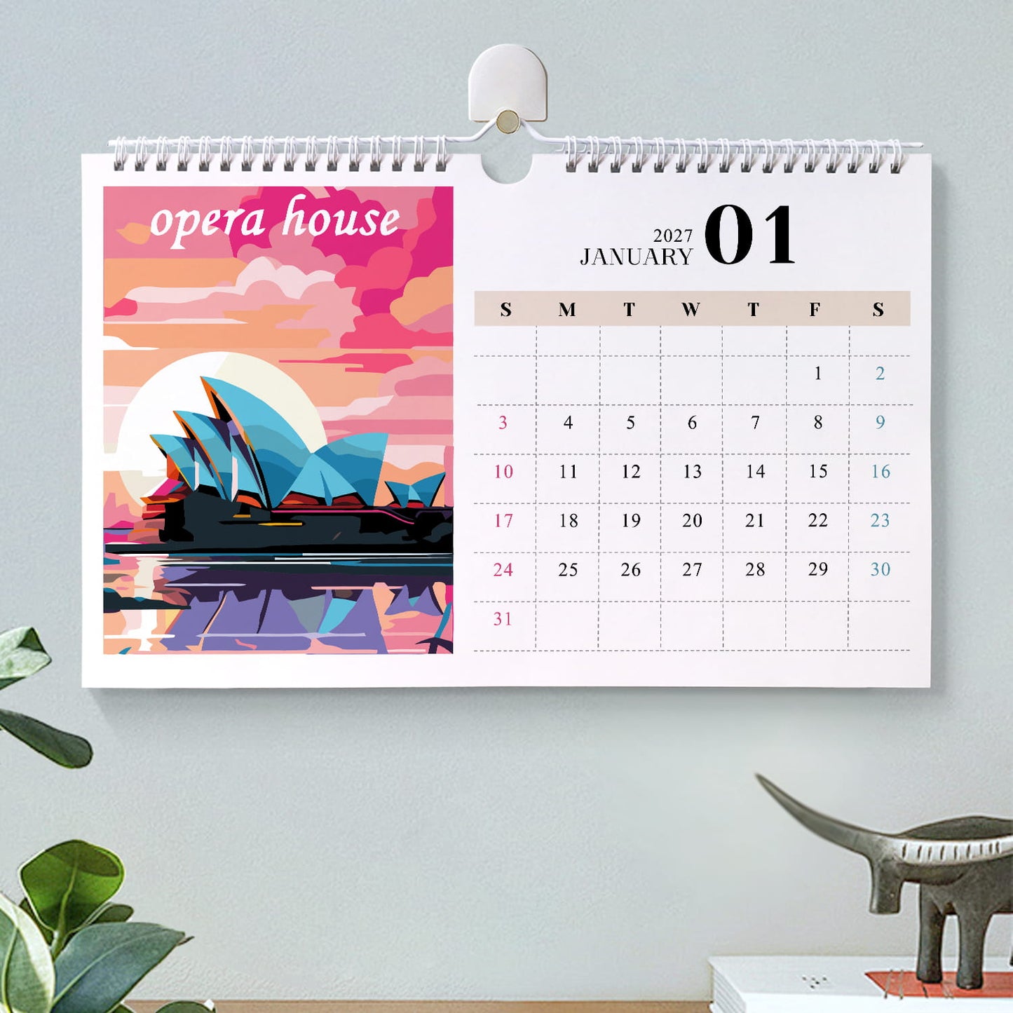 A spiral-bound desk calendar hanging on a light gray wall, showing January 2027 with a colorful Sydney Opera House illustration on the left panel and a weekly date grid on the right. The scene includes a small plant and a dinosaur figurine below.
