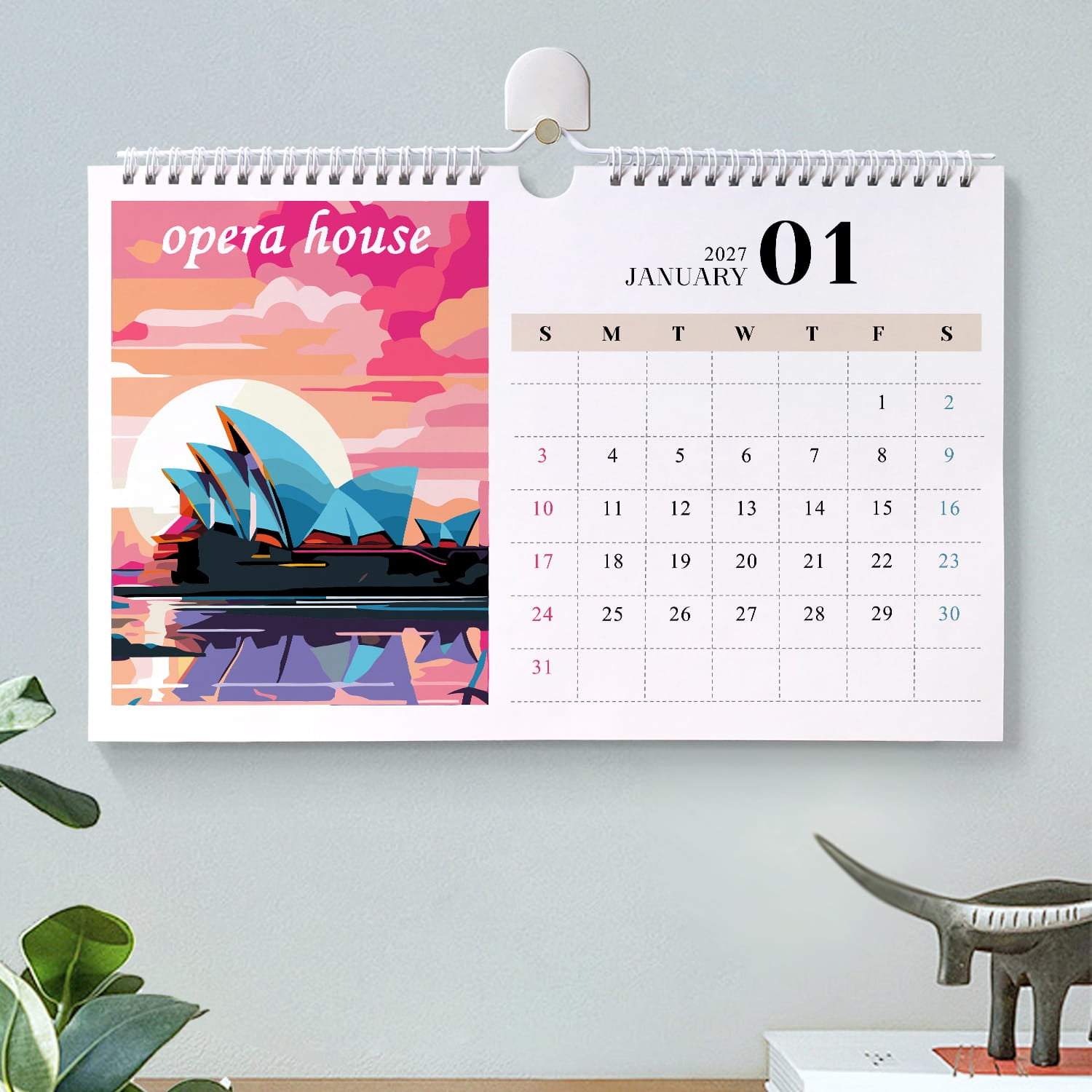 A spiral-bound desk calendar hanging on a light gray wall, showing January 2027 with a colorful Sydney Opera House illustration on the left panel and a weekly date grid on the right. The scene includes a small plant and a dinosaur figurine below.