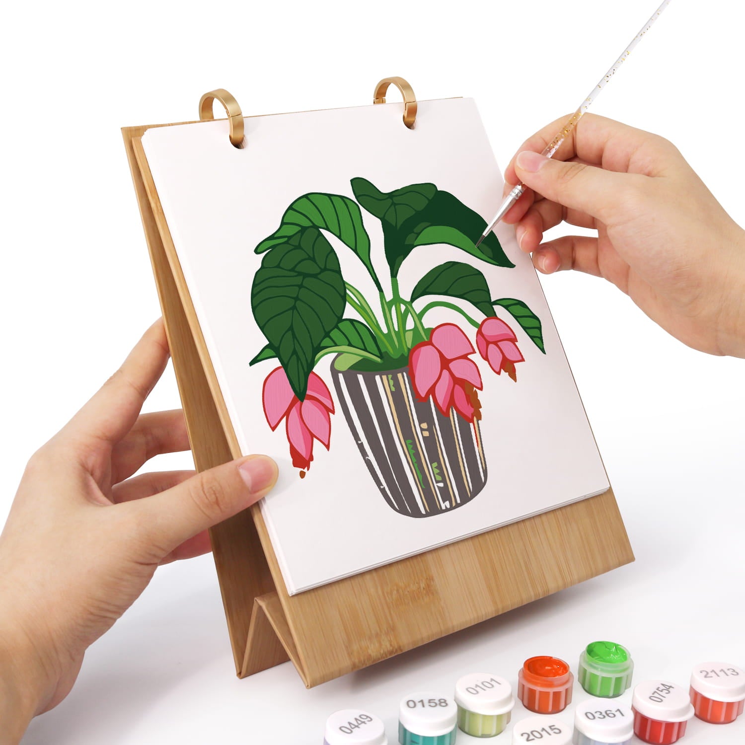 A person's hands are shown painting a floral illustration on a small wooden stand. The scene includes several small paint tubes at the bottom, set against a clean white background.