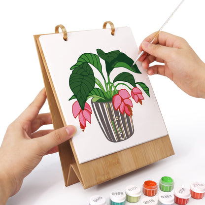 A person's hands are shown painting a floral illustration on a small wooden stand. The scene includes several small paint tubes at the bottom, set against a clean white background.