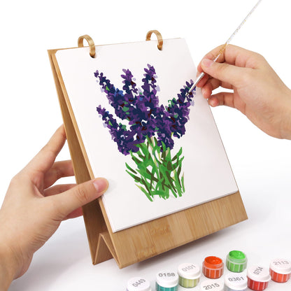 A wooden stand holding a white canvas with a painted purple flower bouquet, shown with two hands painting on it. Small paint tubes are visible at the bottom on a white background.