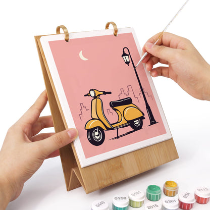 A wooden stand holding a square art canvas with a pink illustration of a yellow scooter, black street lamp, and small crescent moon. Two hands are holding the stand and painting on the canvas with a fine brush.