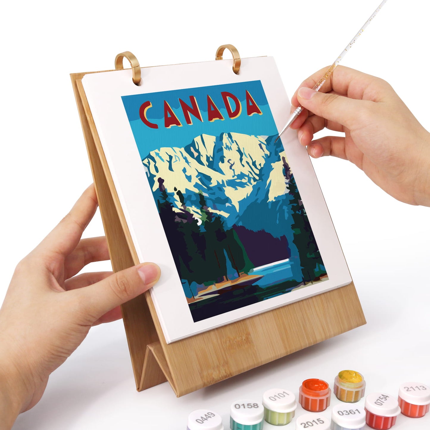 A person's hands are shown painting a landscape art print of Canada on a wooden easel. The print features a blue sky, snowy mountains, and pine trees.