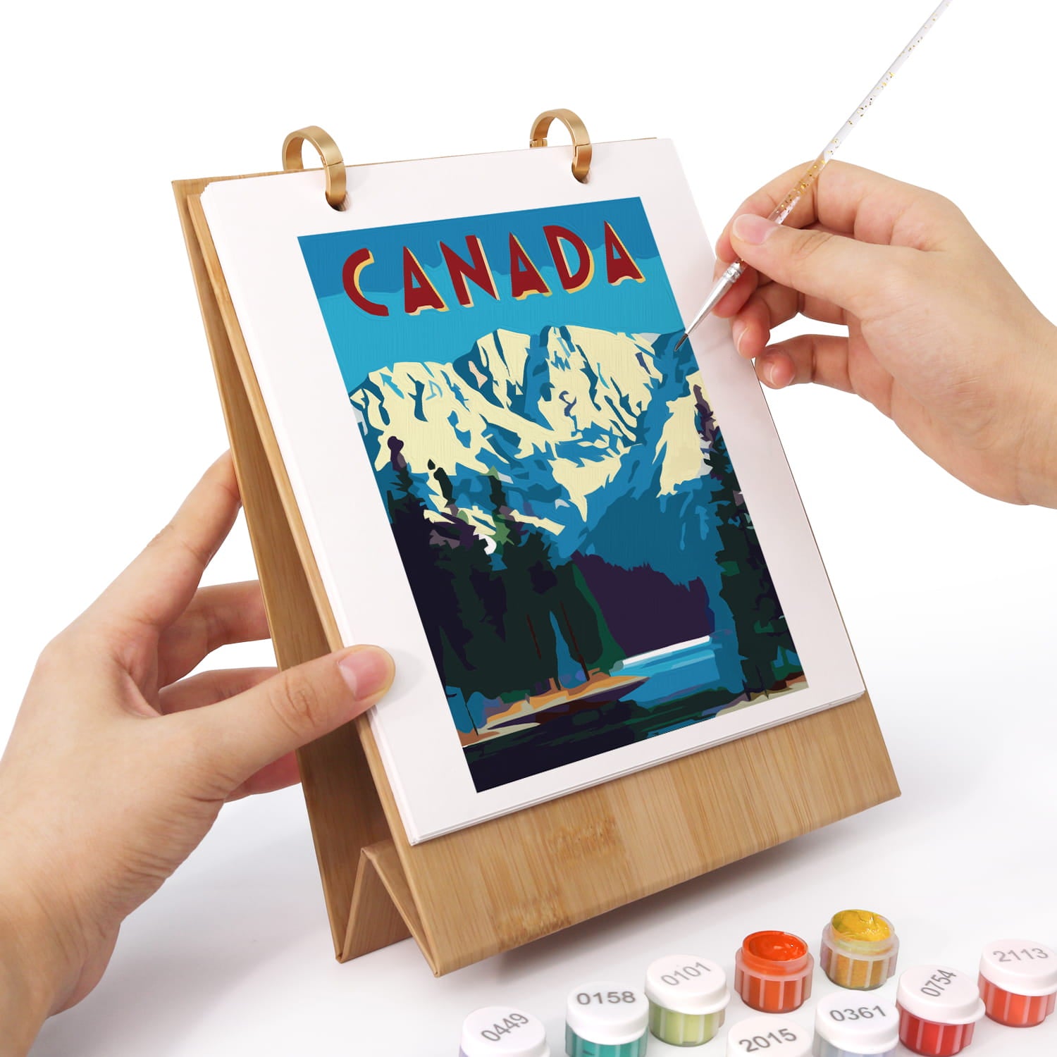 A person's hands are shown painting a landscape art print of Canada on a wooden easel. The print features a blue sky, snowy mountains, and pine trees.