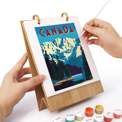 A person's hands are shown painting a landscape art print of Canada on a wooden easel. The print features a blue sky, snowy mountains, and pine trees.
