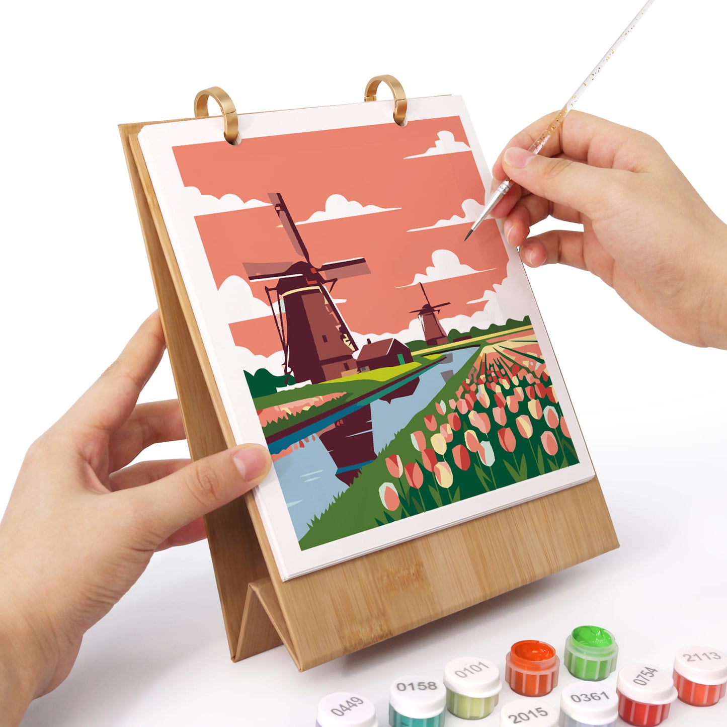 A person holds a bamboo tabletop art frame with a colorful landscape painting of windmills and tulips, while another hand uses a fine brush to apply paint to the canvas. Small paint tubes are visible at the bottom on a white background.