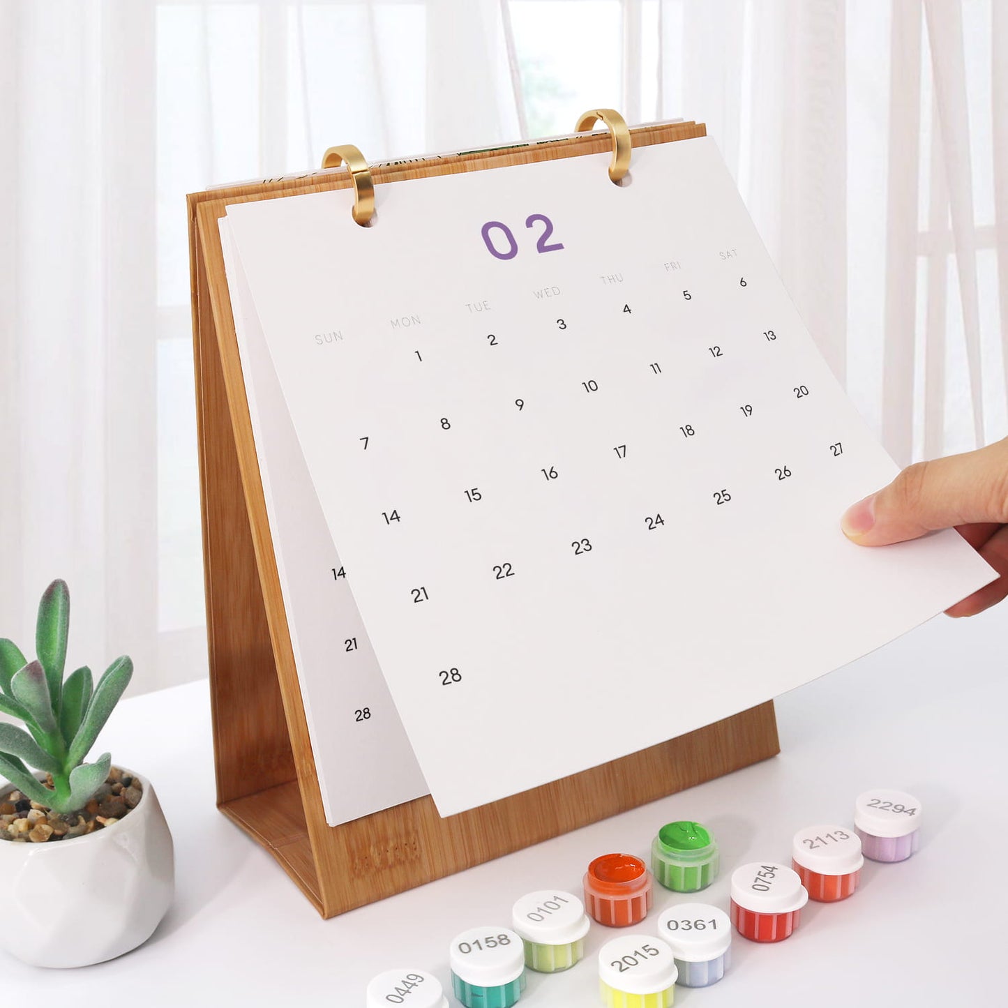 A wooden desktop calendar with a white page showing the number 02 in purple, held open by a hand. Small round decorative caps or lids are lined up in front on a white table, with a small potted succulent and soft window curtains in the background.