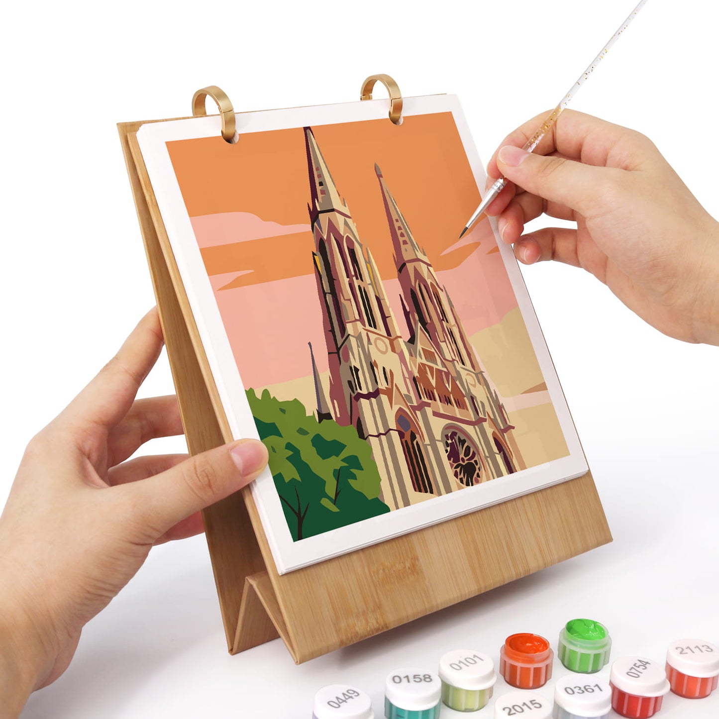 A wooden tabletop art frame holding a colorful cathedral illustration, with a hand using a fine-tip pen to draw on the artwork. Small paint tubes are visible at the bottom on a white background.