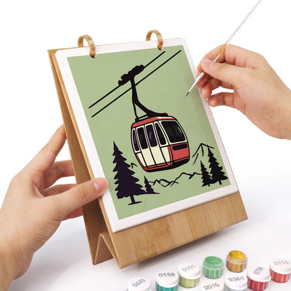 A bamboo stand holding a printed canvas with a cable car illustration, being painted by two hands using a fine brush. Small paint tubes are visible at the bottom on a white background.