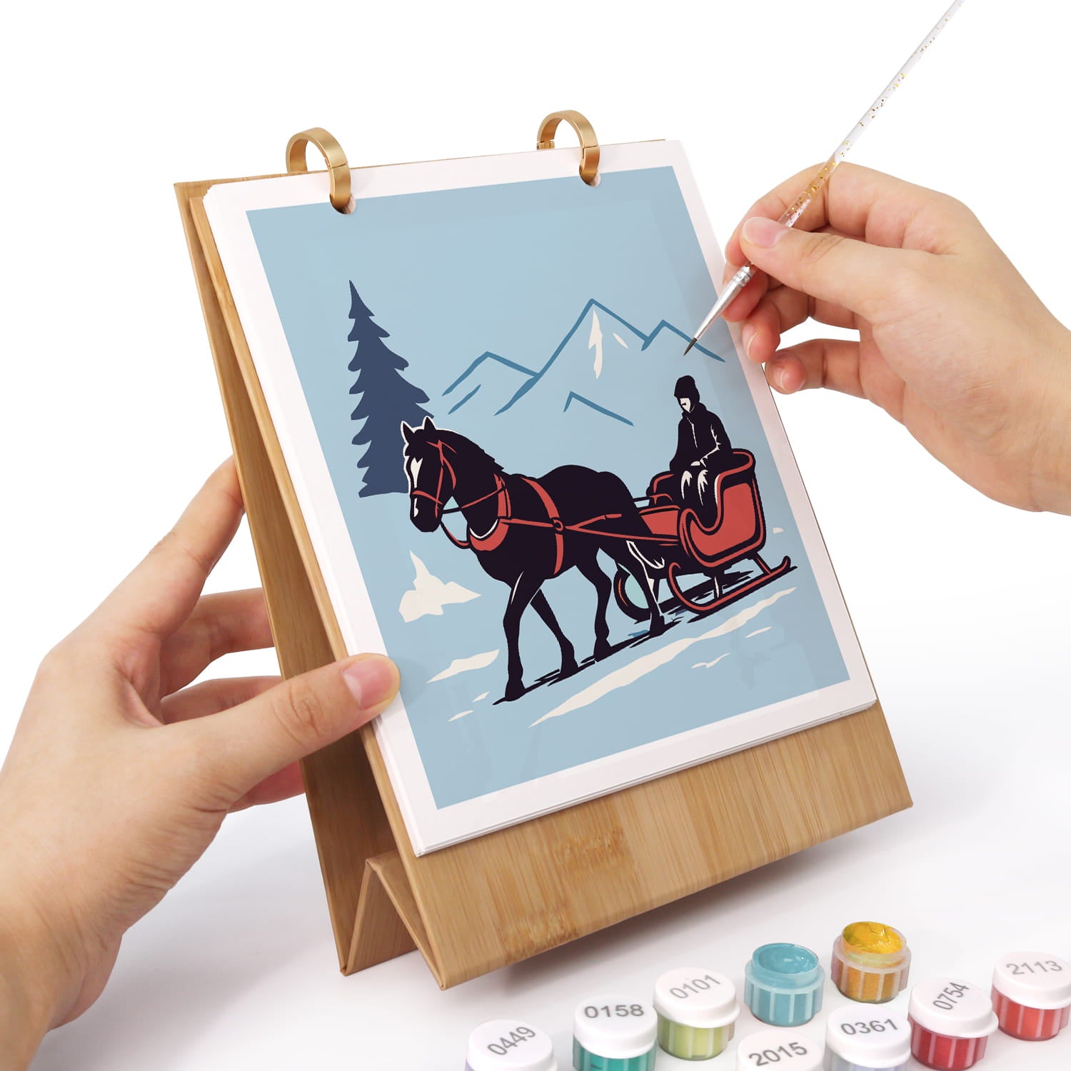 A hands-on painting kit showing a wooden stand holding a canvas with a winter scene illustration of a horse-drawn sleigh. Two hands are visible holding the stand and brush, with small paint pots arranged at the bottom on a white background.