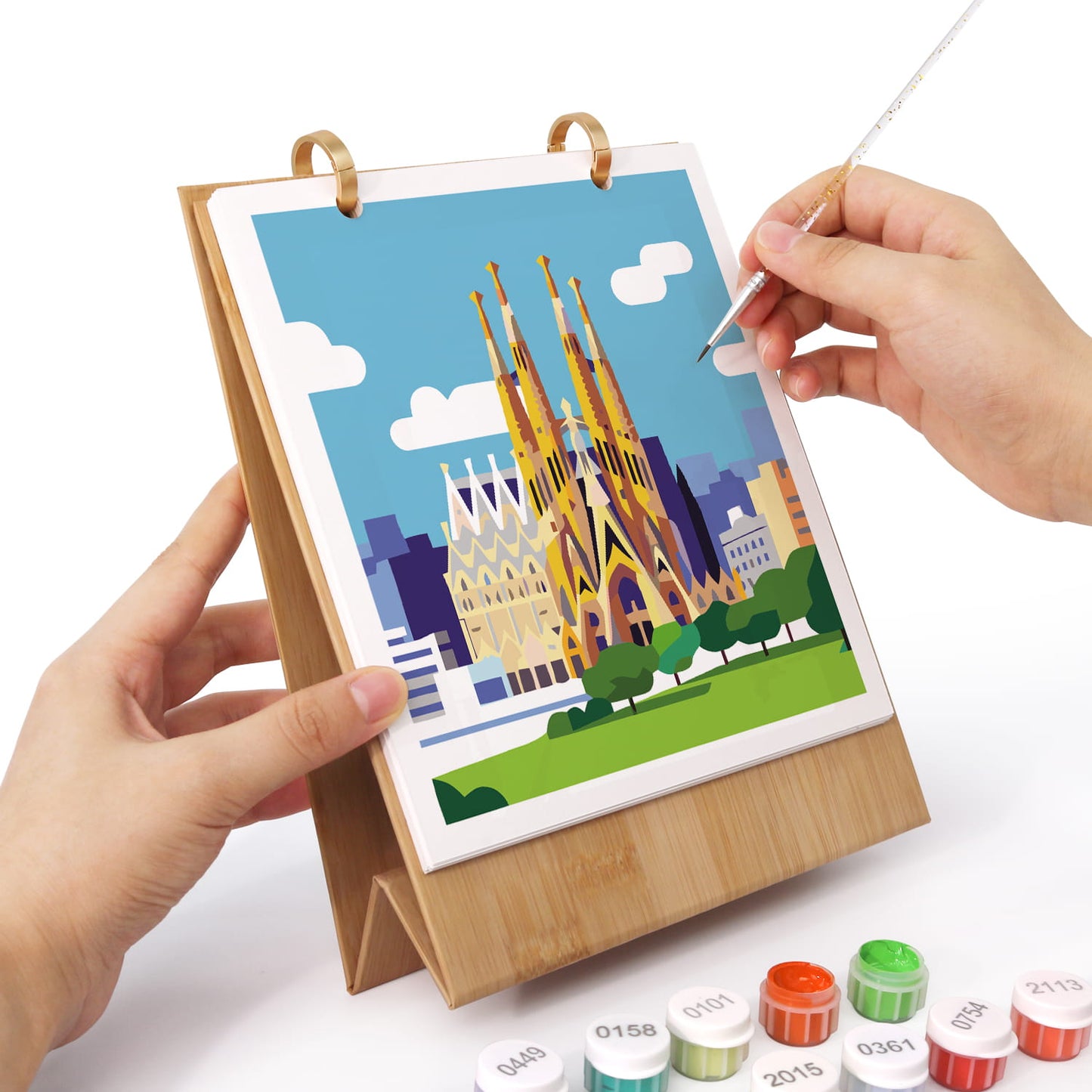 A bamboo tabletop art frame holding a colorful cityscape illustration, with two hands holding it steady while a hand uses a fine-tip pen to draw on the artwork. Small paint tubes are visible at the bottom on a white background.