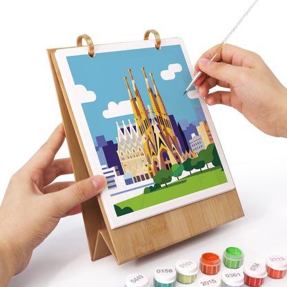 A bamboo tabletop art frame holding a colorful cityscape illustration, with two hands holding it steady while a hand uses a fine-tip pen to draw on the artwork. Small paint tubes are visible at the bottom on a white background.