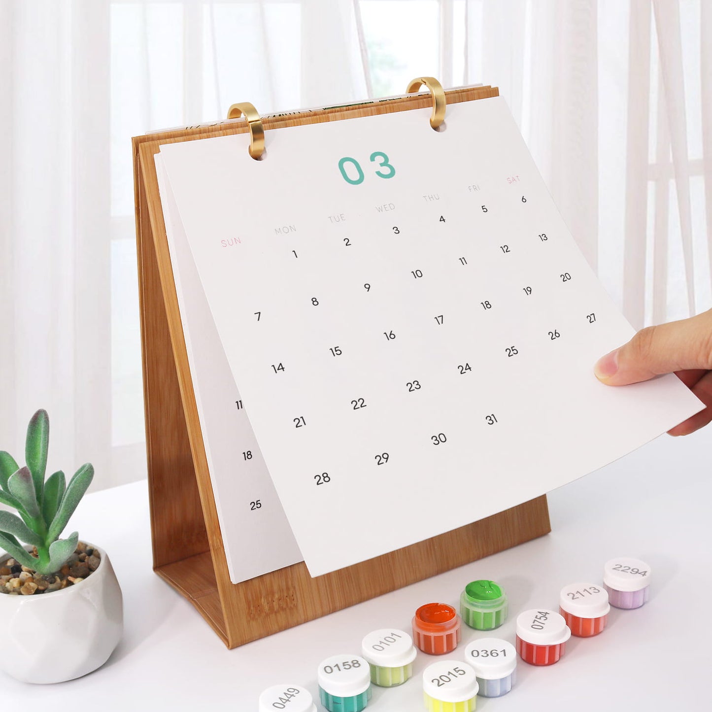 A wooden desktop calendar with a white page showing the number 02 in purple, held open by a hand. Small round decorative caps or lids are lined up in front on a white table, with a small potted succulent and soft window curtains in the background.
