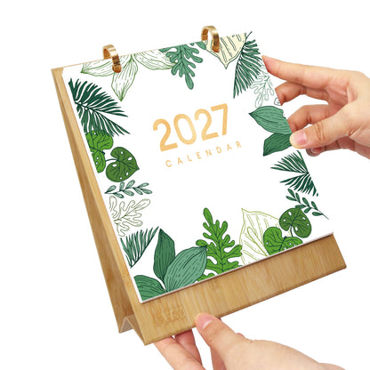 A 2027 desk calendar with a bamboo base and gold wire rings, featuring a white cover decorated with green tropical leaves and gold text. Two hands are shown holding the calendar against a plain white background.