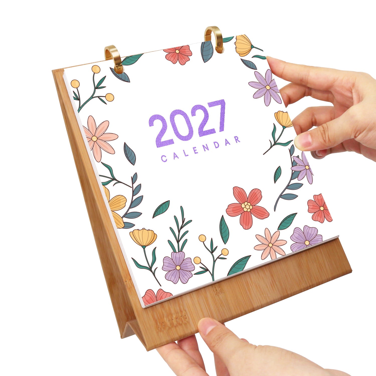 A 2027 desk calendar with a bamboo stand, held by two hands against a white background. The calendar cover is white with a colorful floral border and purple text reading '2027 CALENDAR'.