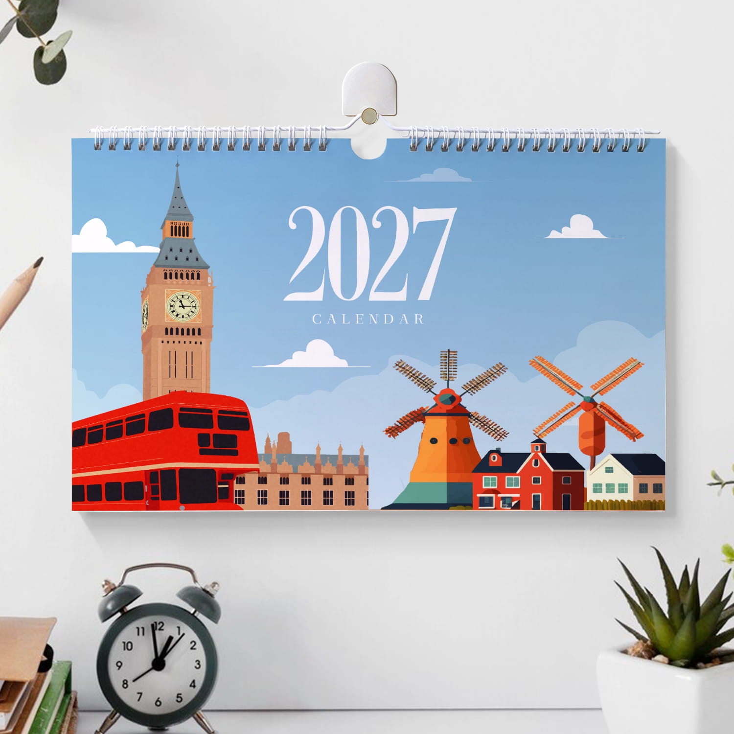 A 2027 wall calendar featuring a London-themed illustration with Big Ben, red double-decker buses, windmills, and red houses under a blue sky, displayed on a white wall above a desk with an alarm clock and small plant.