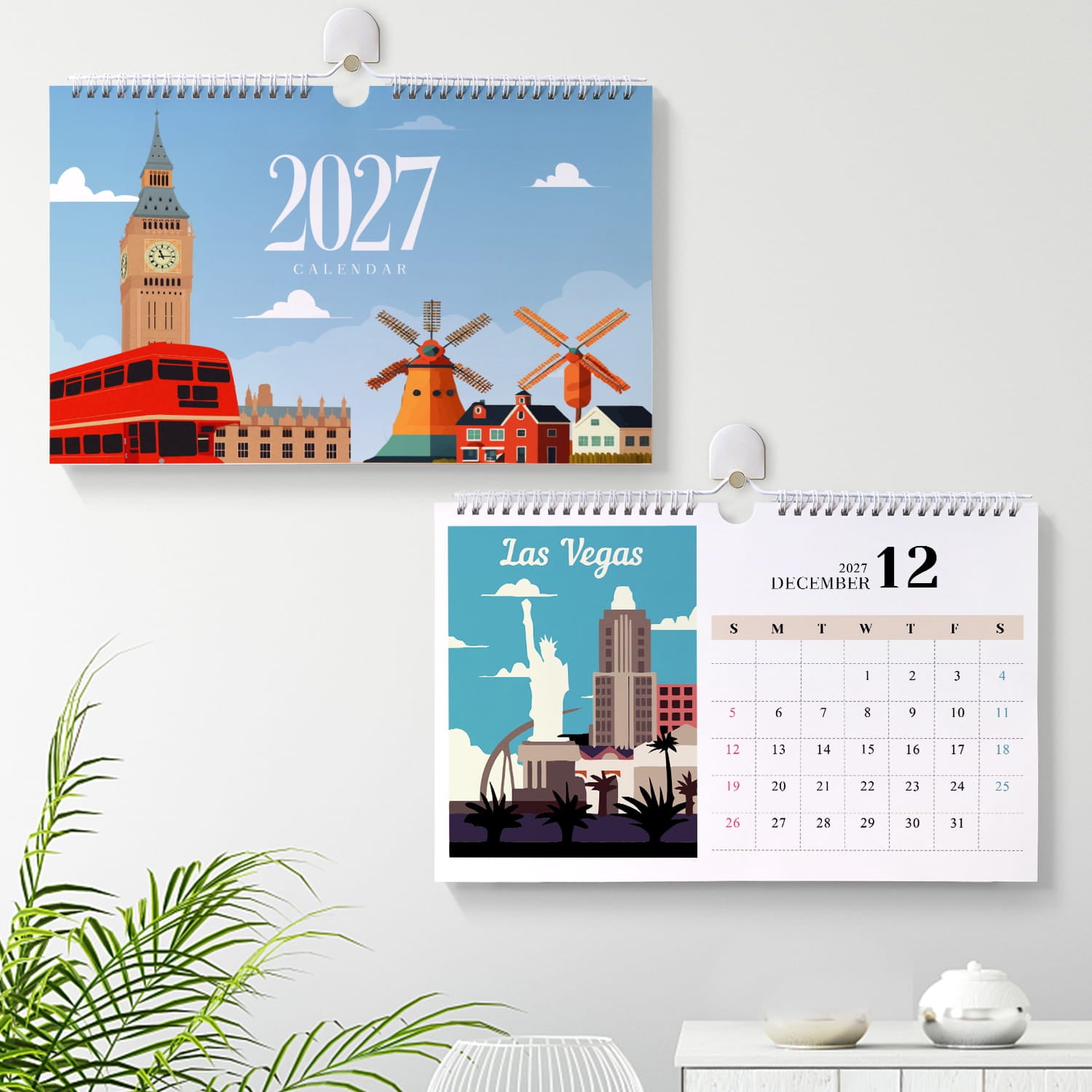 Two spiral-bound wall calendars hanging on a light gray wall, one showing a 2027 London calendar and the other a Las Vegas December 12 calendar. A green plant and a small table with a teapot are visible in the foreground.