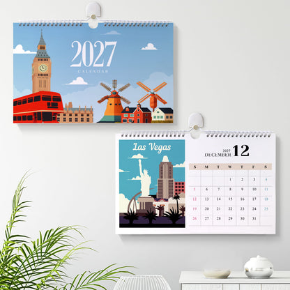Two spiral-bound wall calendars hanging on a light gray wall, one showing a 2027 London calendar and the other a Las Vegas December 12 calendar. A green plant and a small table with a teapot are visible in the foreground.