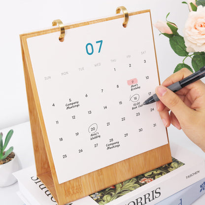 Calendar with wooden stand on a magazine, hand holding a pen, and flowers in the background