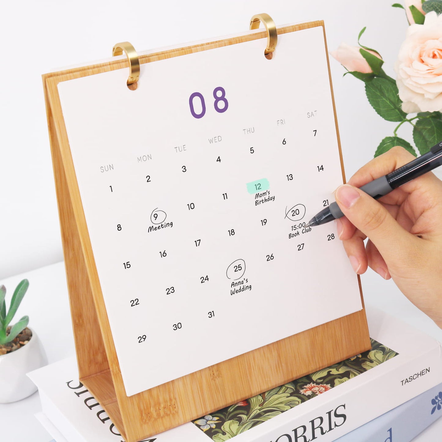 A bamboo desk calendar with a white monthly calendar page, held by a hand with a black pen. The page shows the number 08 in purple and various event markers.