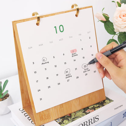 A bamboo desk calendar with a white page showing the 10th and a hand writing on it, styled on books with a rose in the background.
