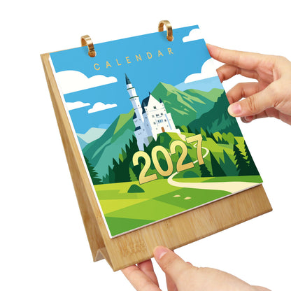 A bamboo desk calendar with a 2027 cover featuring a colorful illustration of Neuschwanstein Castle, held by two hands against a white background.