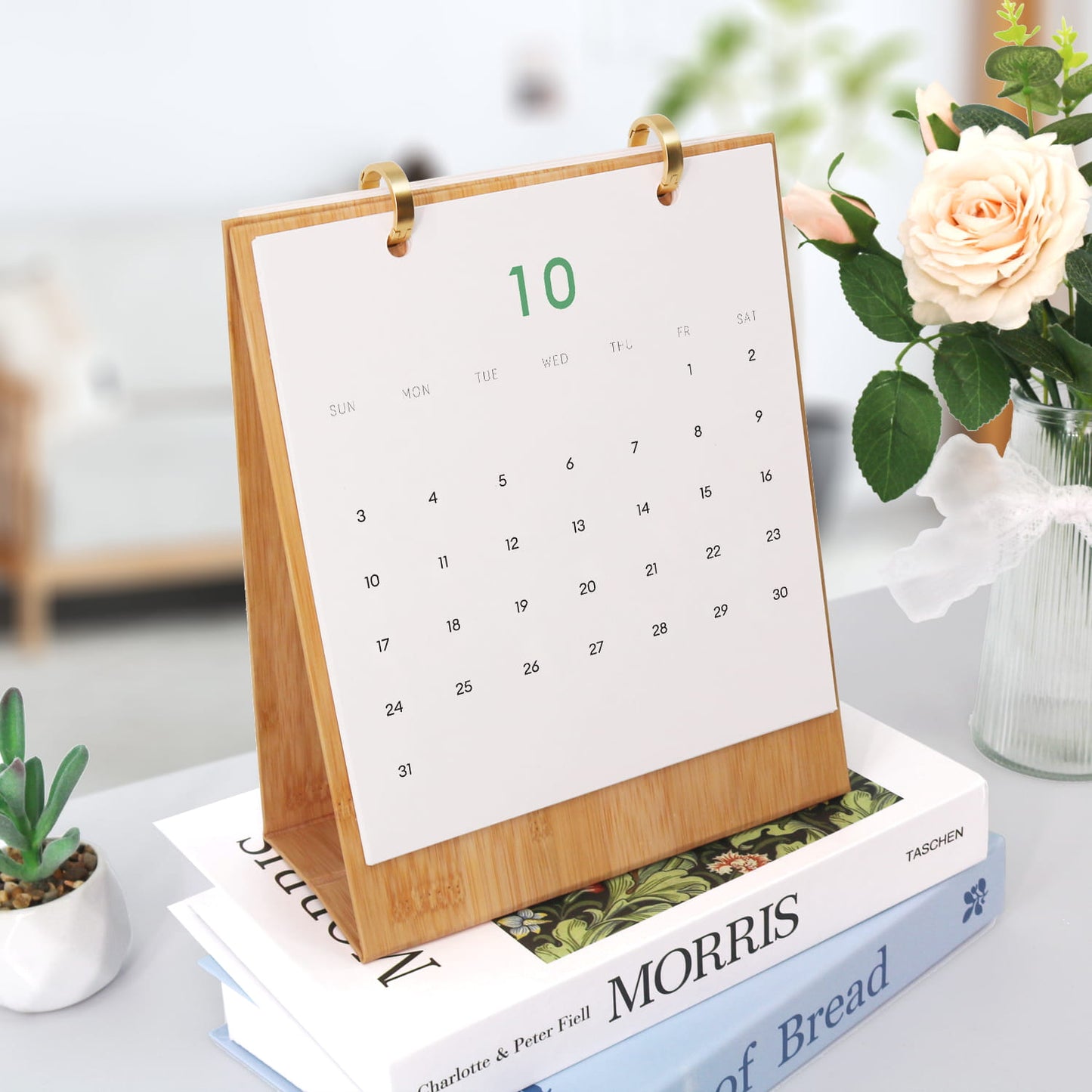 A bamboo desk calendar with a white page showing the 10th highlighted in green, styled on top of books with a small plant and flowers in the background.