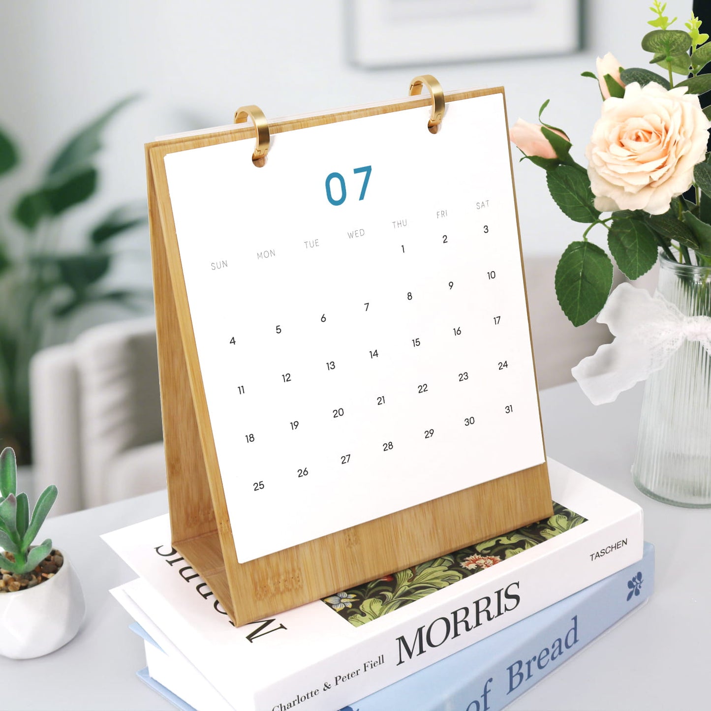 A wooden desktop calendar standing on books, with a white page showing the 7th in blue, styled on a table with flowers and plants in a bright indoor setting.