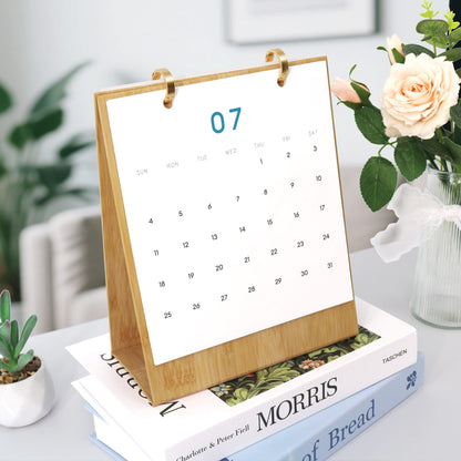 A wooden desktop calendar standing on books, with a white page showing the 7th in blue, styled on a table with flowers and plants in a bright indoor setting.