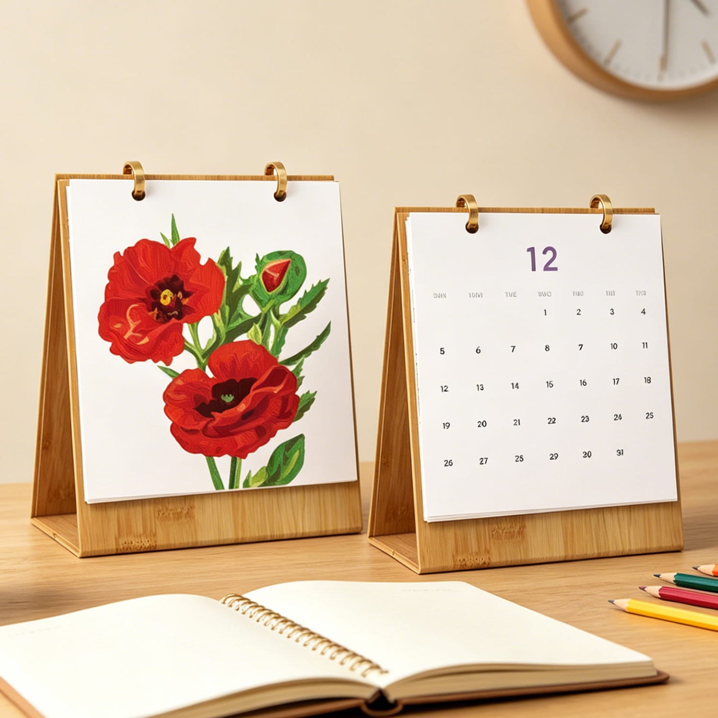 A bamboo desktop calendar stand on a wooden table, showing a floral page with red poppy flowers on the left and the number 12 on the right. An open notebook and colored pencils are in the foreground, with a wall clock blurred in the background.