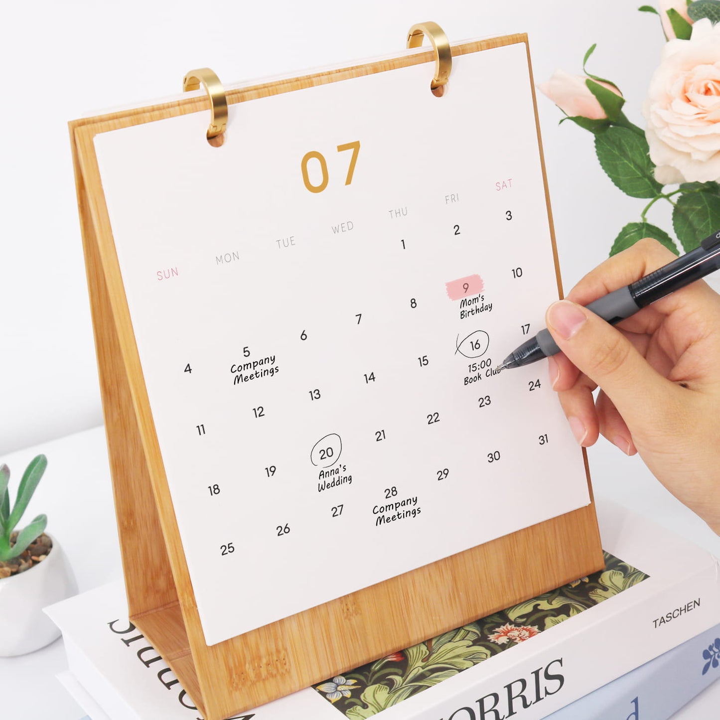 A bamboo desktop calendar with a white page showing the 7th and a hand writing with a black pen, styled on books with flowers in the background.