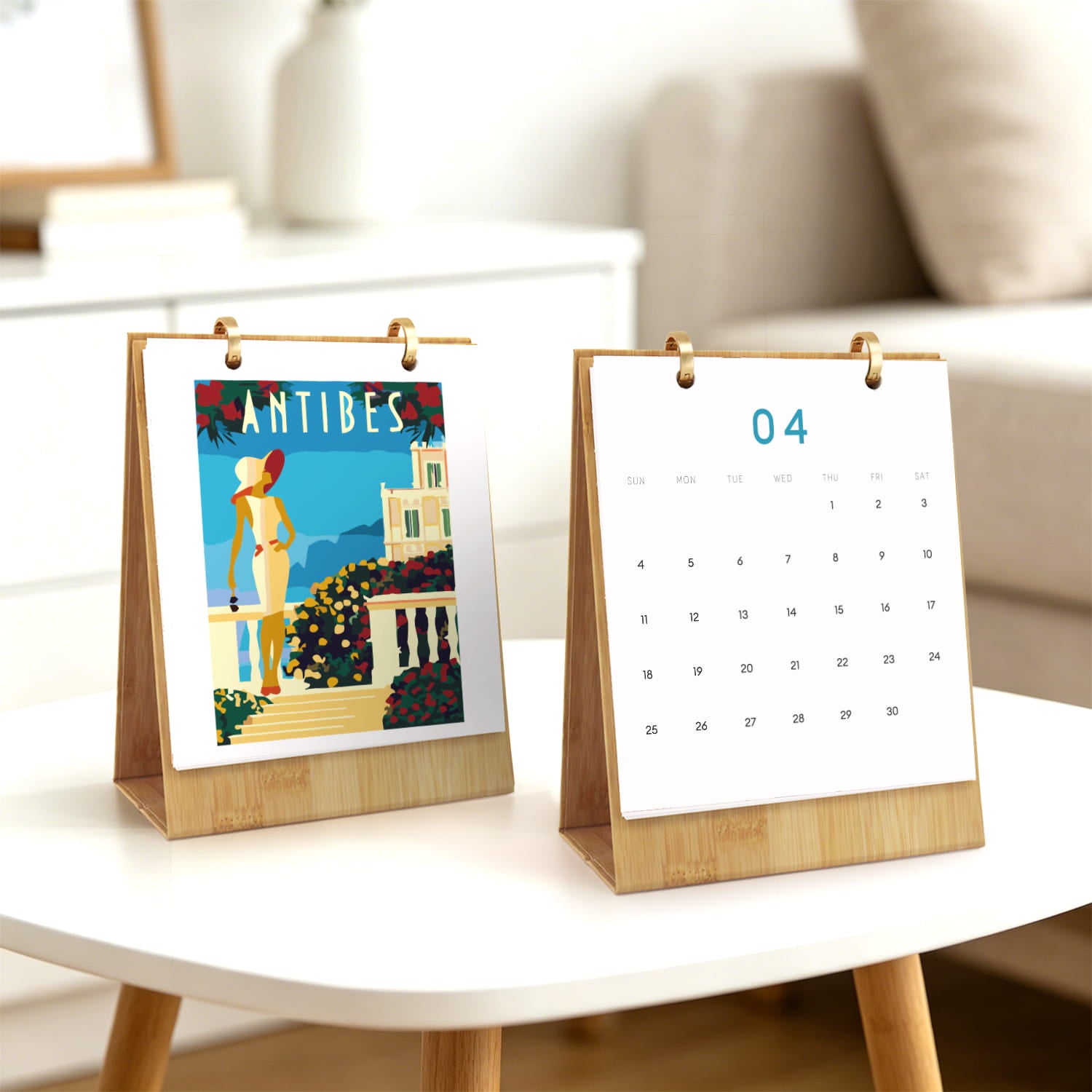 Two small wooden desktop calendar stands on a white table in a bright living room. One stand shows a colorful Antibes travel poster, and the other shows the date 04 with a blank calendar grid.