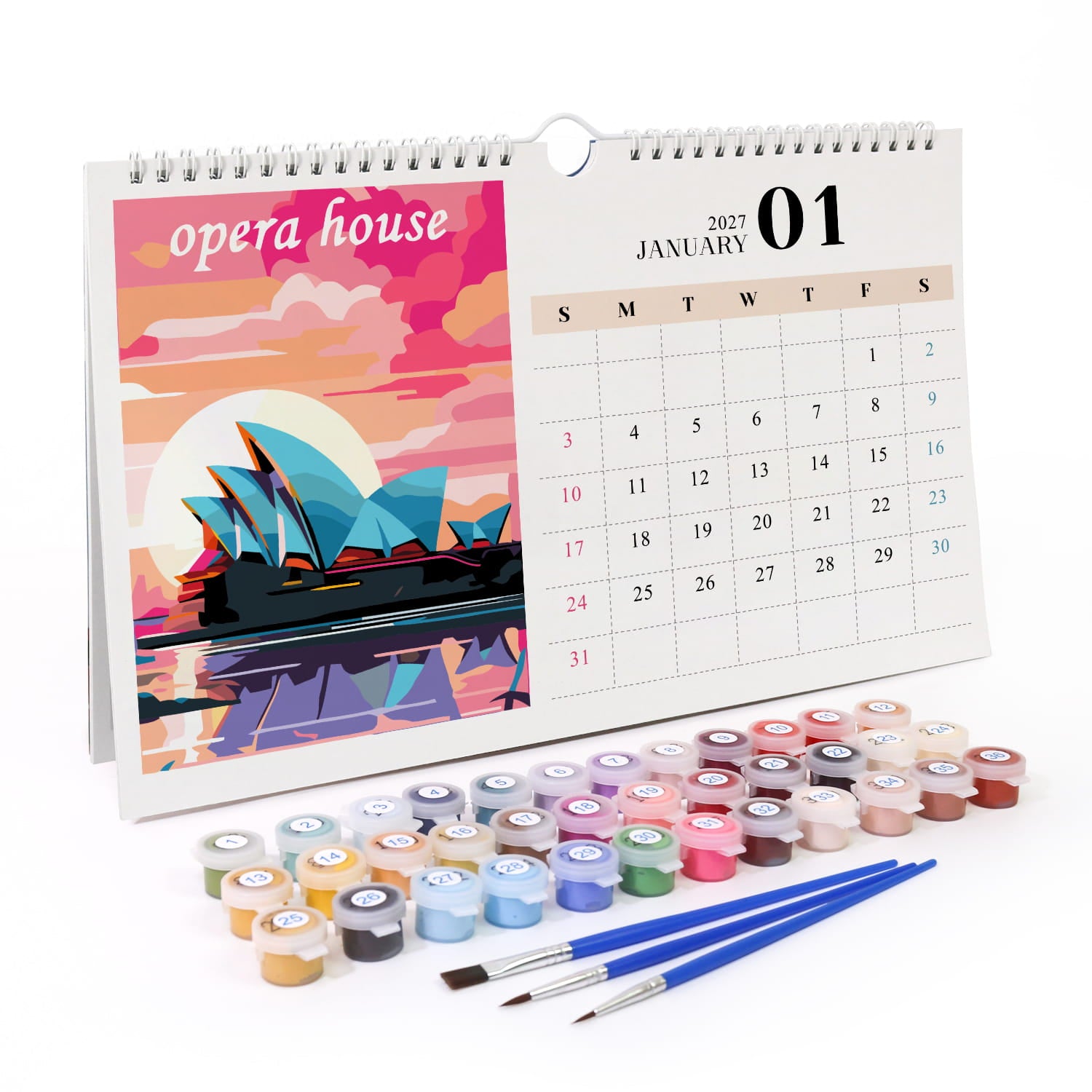 A spiral-bound desk calendar showing January 1st with an illustrated Opera House scene, displayed with multiple small paint pots and three blue-handled brushes on a white background.