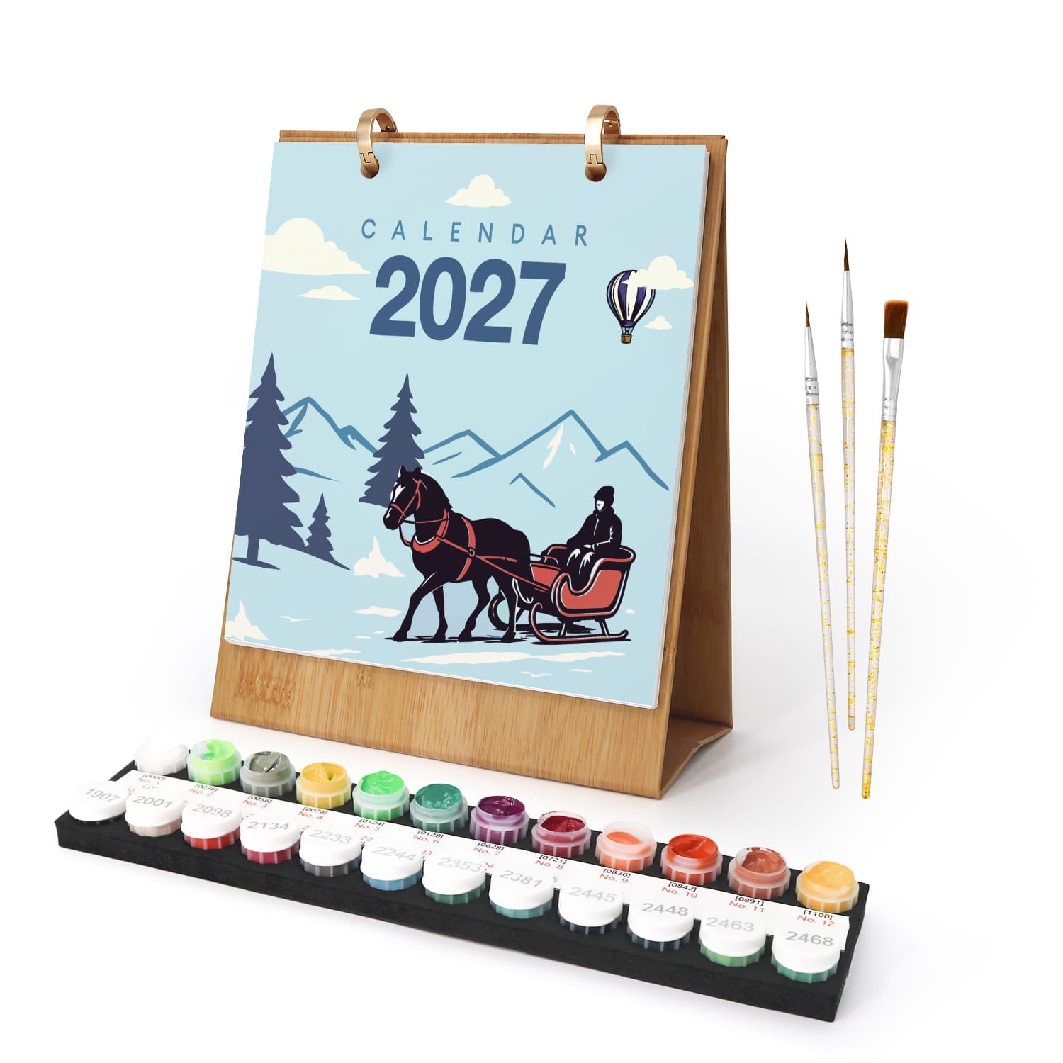 A 2027 calendar with a winter illustration, shown standing on a wooden base with a set of paint pots and three paintbrushes on a white background.