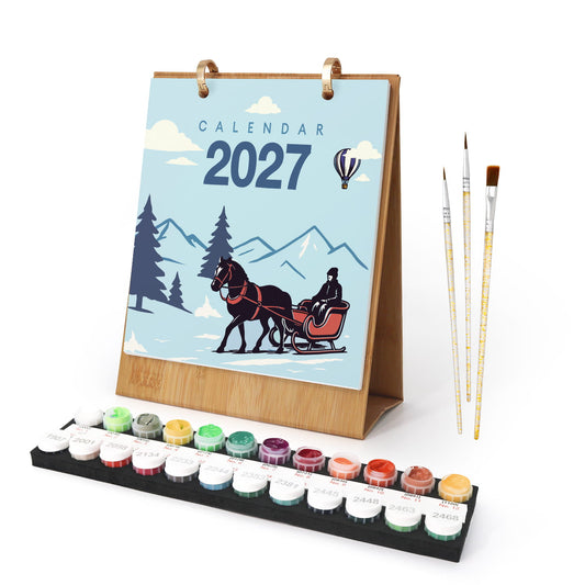 A 2027 calendar with a winter illustration, shown standing on a wooden base with a set of paint pots and three paintbrushes on a white background.