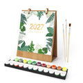 A 2027 desk calendar with a tropical leaf pattern and gold text, shown with a wooden stand, a set of paint pots, and three paintbrushes on a white background.