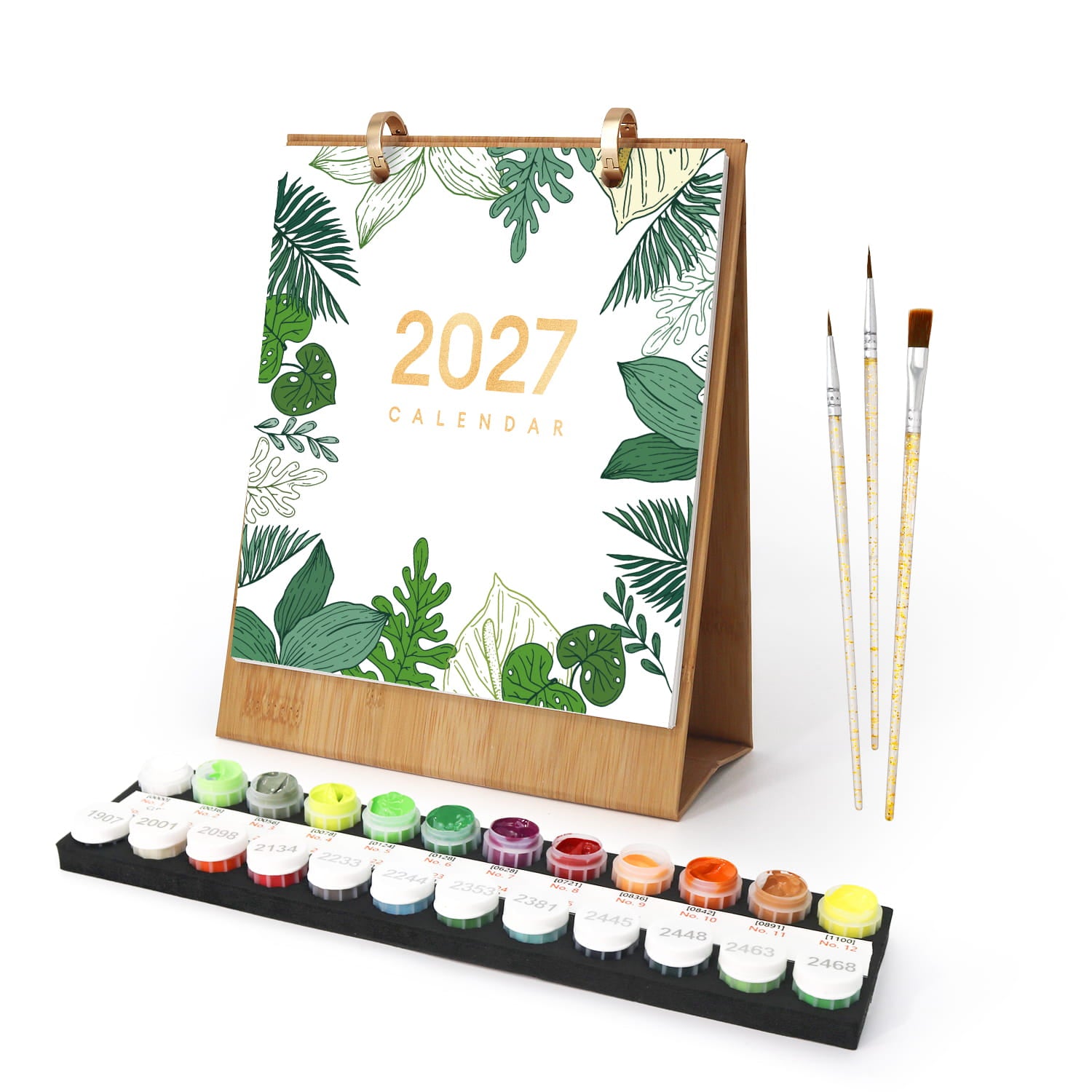 A 2027 desk calendar with a tropical leaf pattern and gold text, shown with a wooden stand, a set of paint pots, and three paintbrushes on a white background.