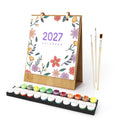 A 2027 desk calendar with a floral border design, shown with a set of paint brushes and a tray of colorful paint pots on a white background.