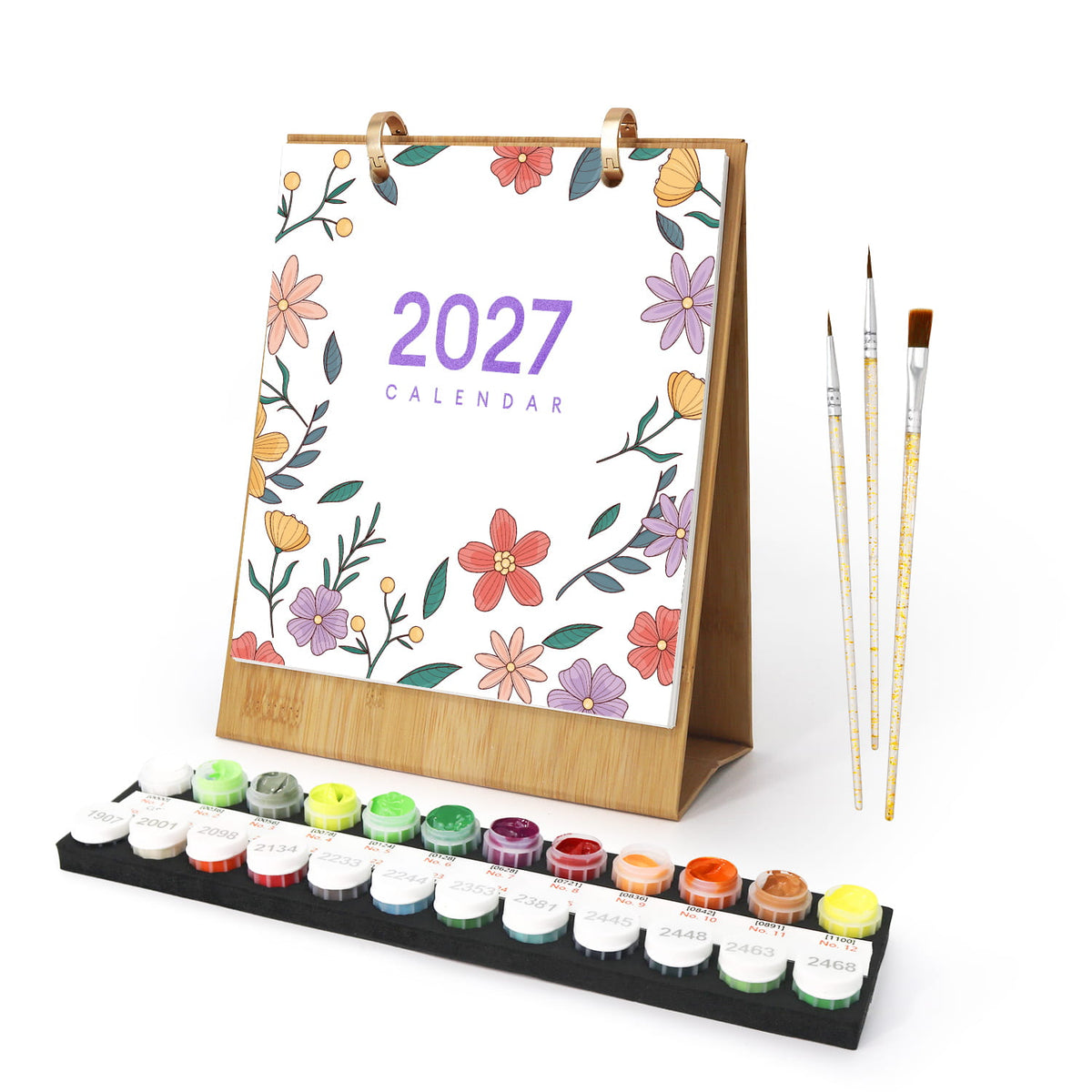 A 2027 desk calendar with a floral border design, shown with a set of paint brushes and a tray of colorful paint pots on a white background.