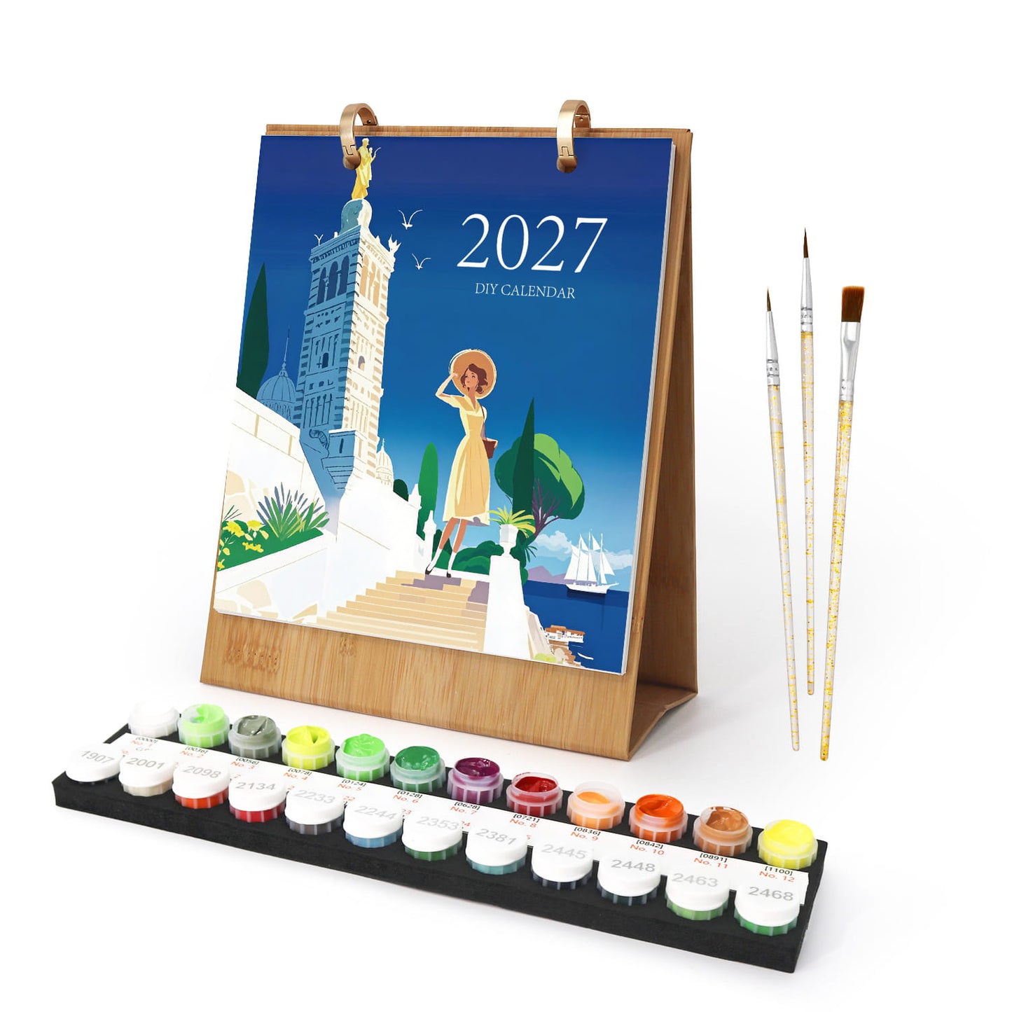 A 2027 DIY art calendar set featuring a wooden-framed stand with a colorful illustrated cover, a row of small paint pots on a black tray, and three paintbrushes on a white background.