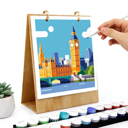 A tabletop canvas painting set with a wooden easel holding a colorful London skyline illustration, shown with a hand holding a paintbrush and a row of paint tubes in the foreground on a white background.