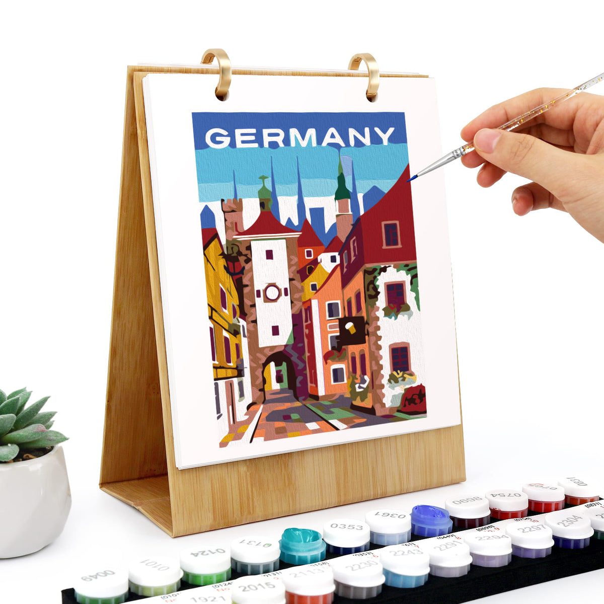 
A wooden-framed diamond painting canvas featuring a colorful Germany cityscape with the word 'GERMANY' at the top. A hand holds a fine paintbrush over the artwork, with paint pots arranged along the bottom and a small potted succulent on the left.