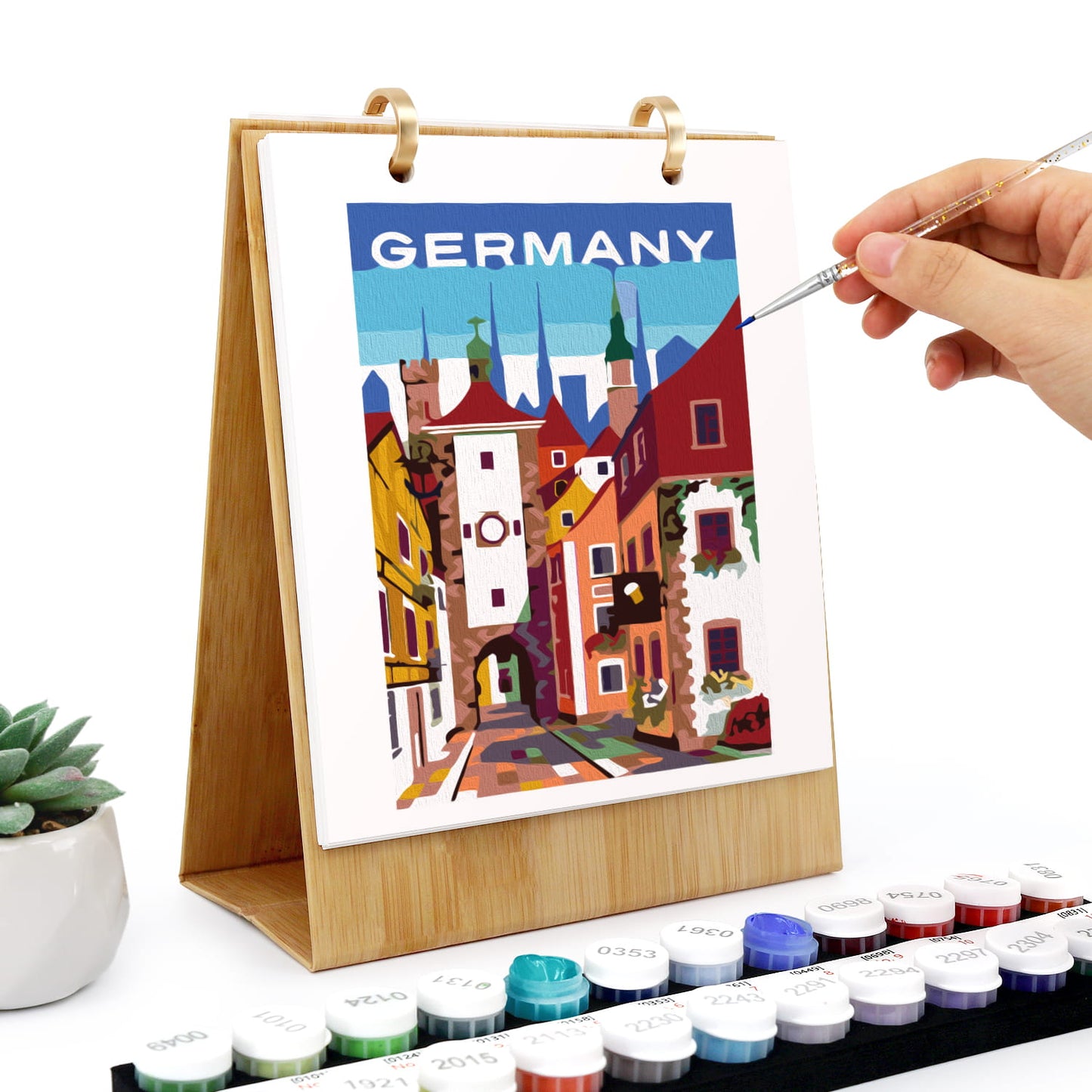 A wooden-framed diamond painting canvas featuring a colorful Germany cityscape with the word 'GERMANY' at the top. A hand holds a fine paintbrush over the artwork, with paint pots arranged along the bottom and a small potted succulent on the left.