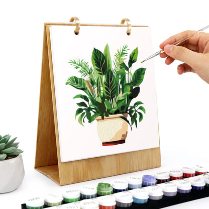 A wooden tabletop canvas painting set with a plant in a pot, shown with a hand holding a paintbrush and paint pots in the foreground on a white background.
