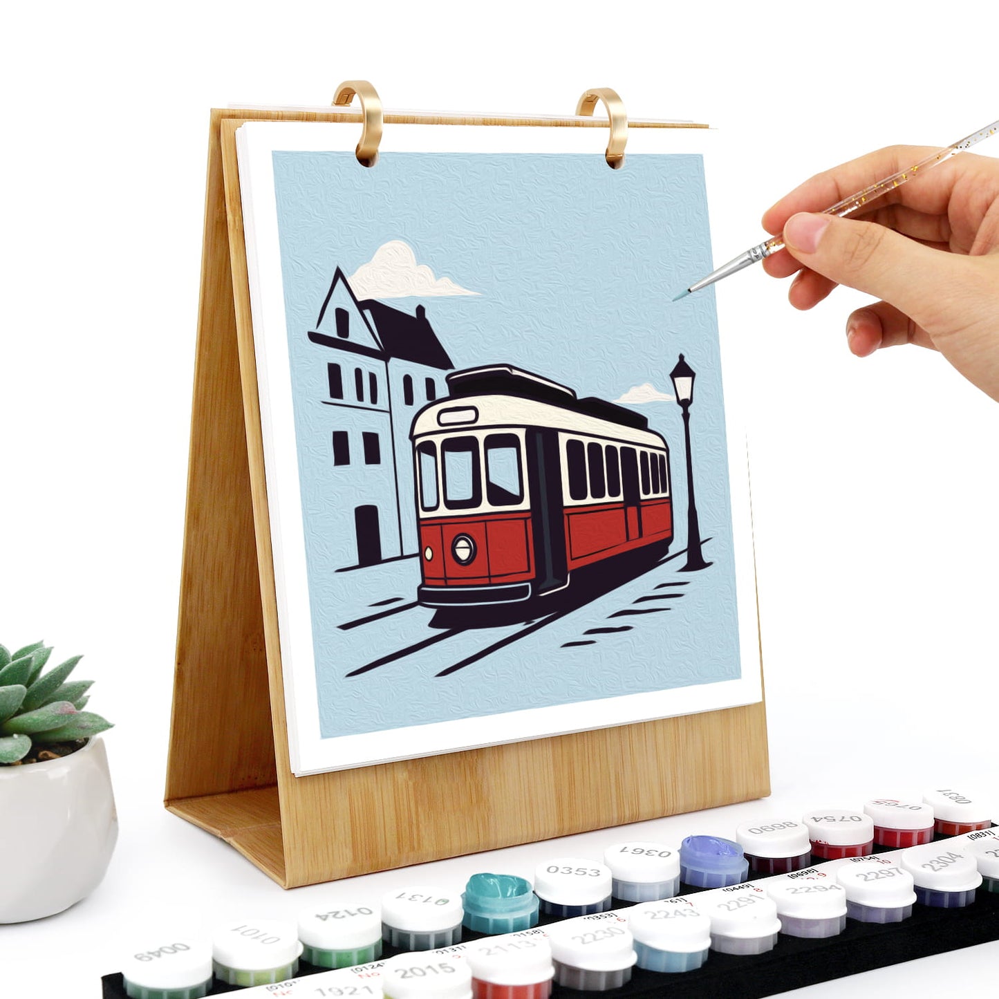 A wooden stand holding a colorful city scene painting with a red tram, white clouds, and buildings. A hand holds a fine paint brush near the artwork, with small round paint pots arranged along the bottom and a small potted succulent on the left.